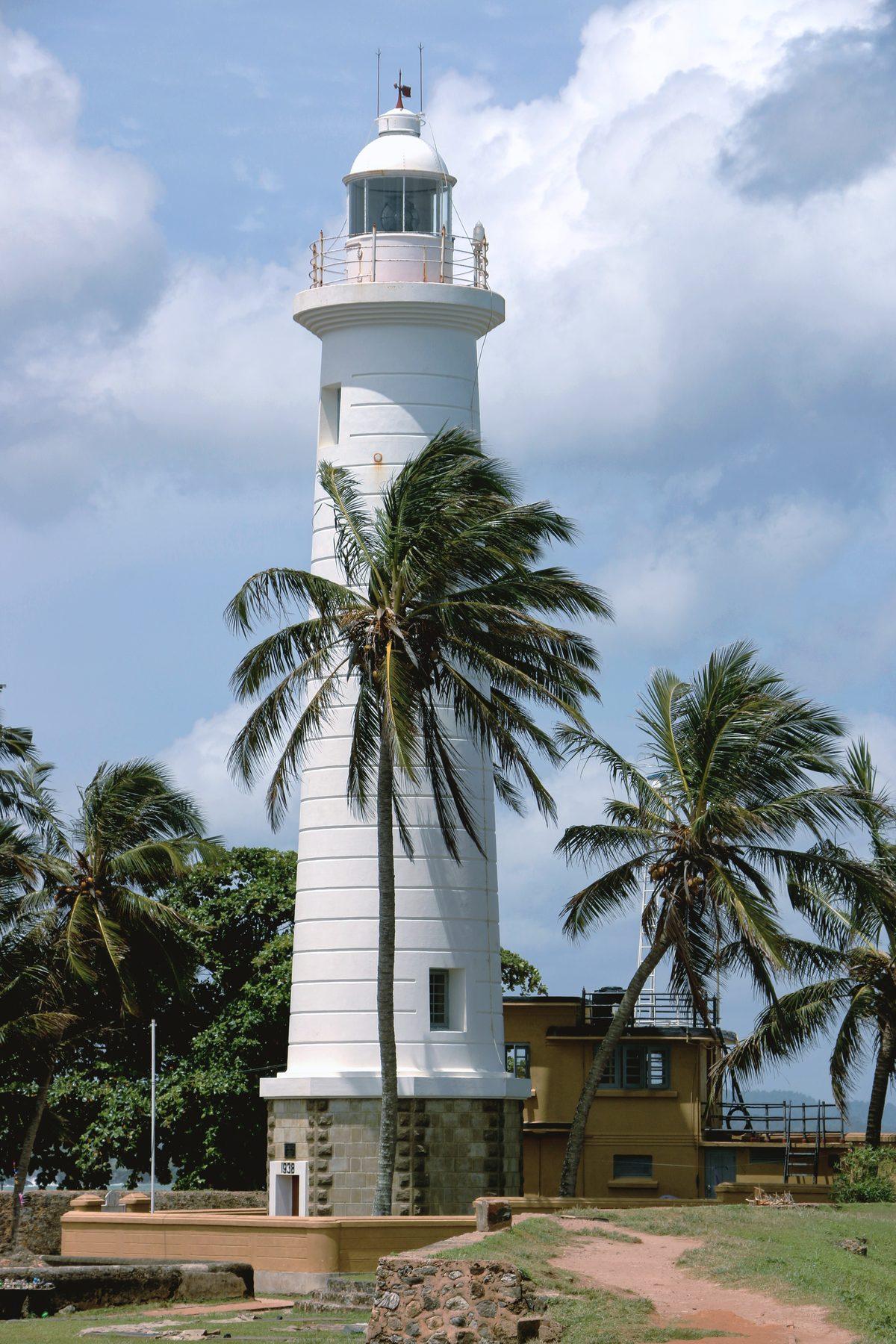A white lighthouse rising above palm trees under a blue sky